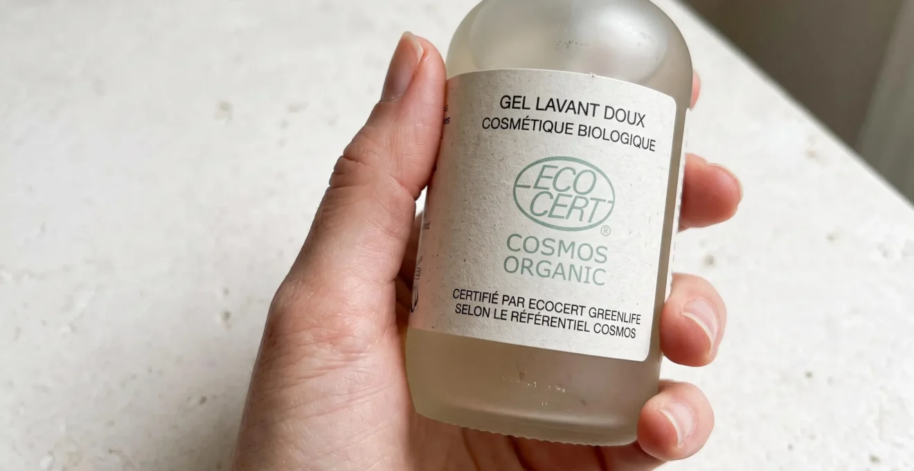 Gros plan sur un flacon de cosmétique bio pour enfant avec logo de certification Ecocert visible, tenu par une main adulte sur fond neutre blanc
